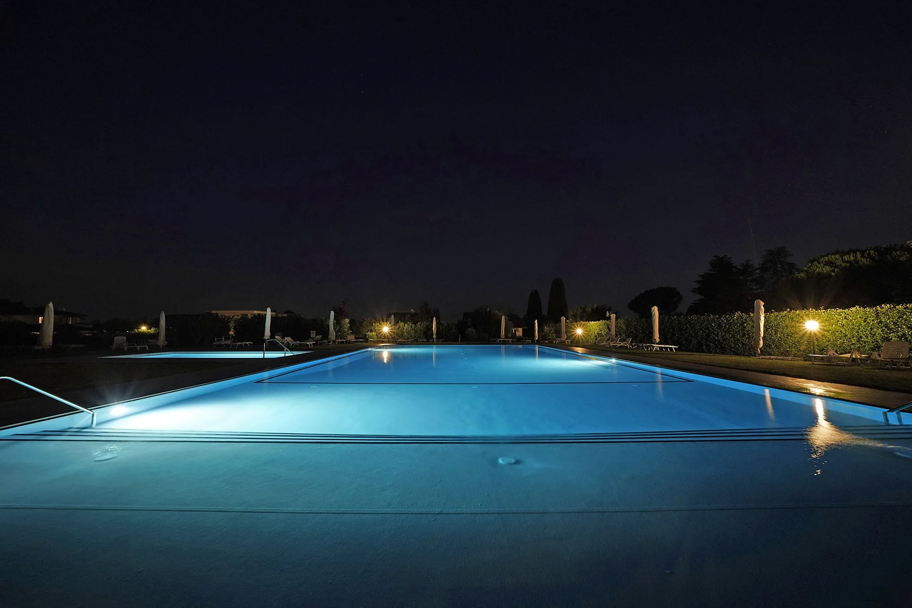 An illuminated swimming pool at night reflects blue light beneath a dark sky.