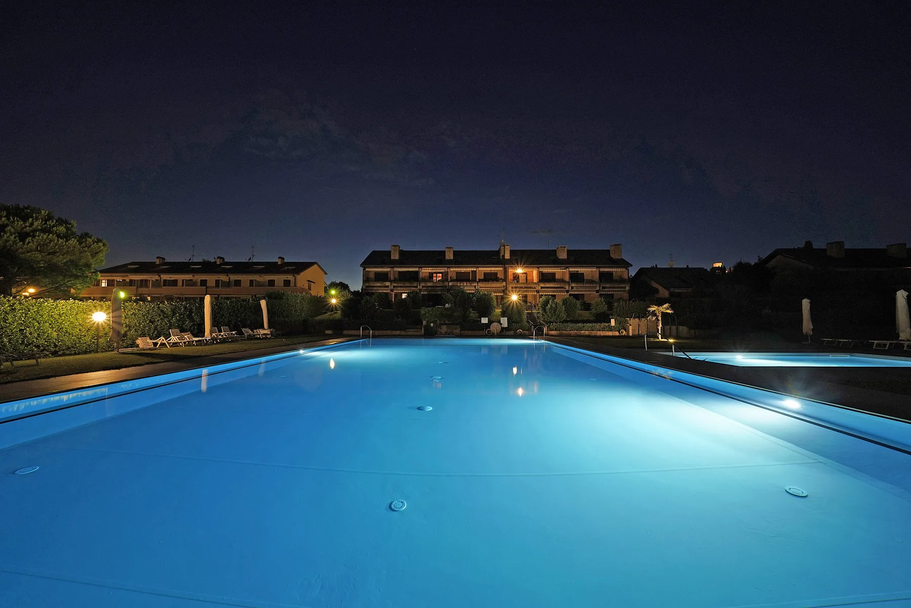 A swimming pool glows blue at night, with buildings and landscaped grounds beyond.