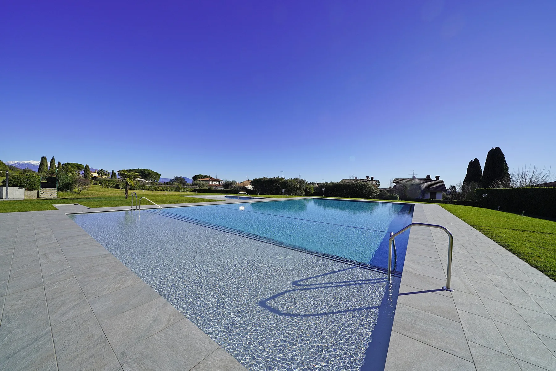 An outdoor swimming pool is surrounded by a broad paved deck and green lawn.