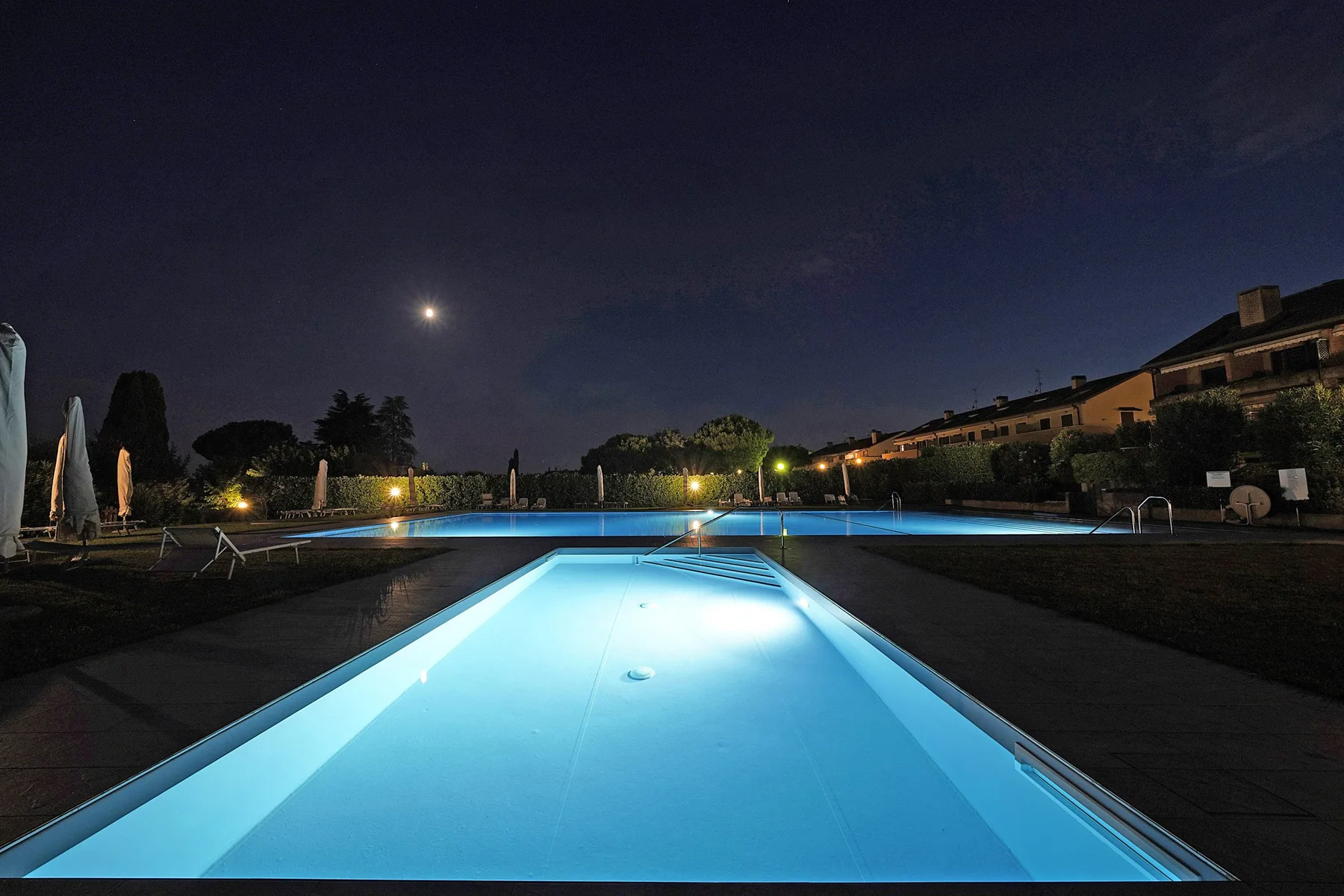 A lit outdoor swimming pool at night, with buildings and trees in the background.
