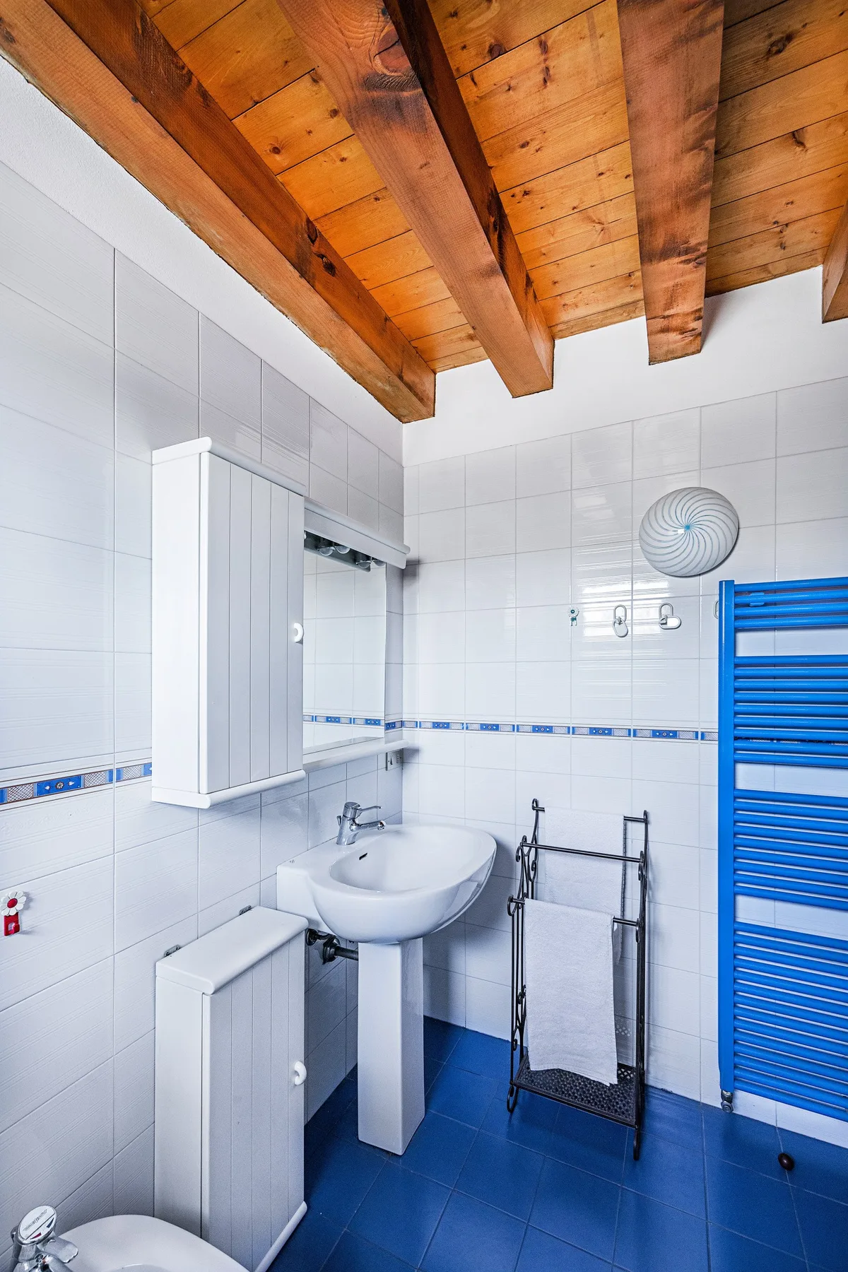 A tiled bathroom with a pedestal sink, blue floor, and wooden ceiling beams.