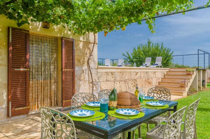 An outdoor dining area sits beneath leafy vines beside a stone building.