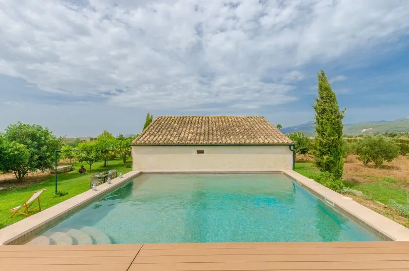 An outdoor swimming pool sits beside a tiled-roof building amid green countryside.