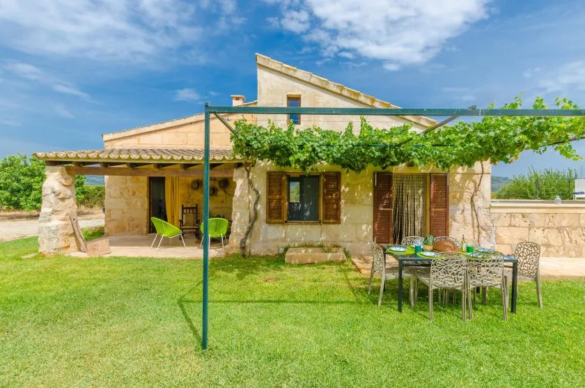 A stone house with a vine-covered pergola over a grassy outdoor dining area.