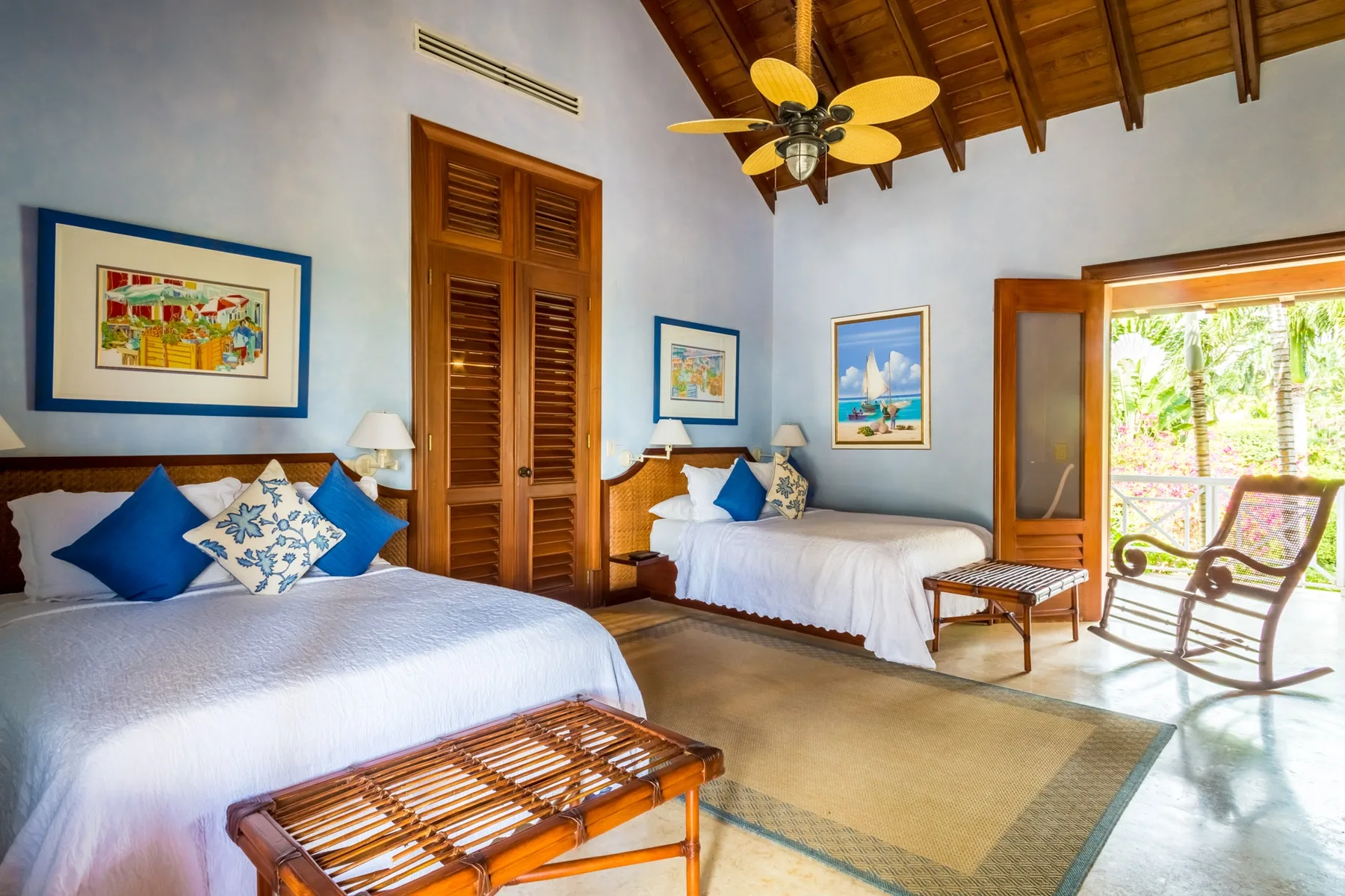 A bright bedroom with two beds, framed artwork, and an open doorway to a patio.