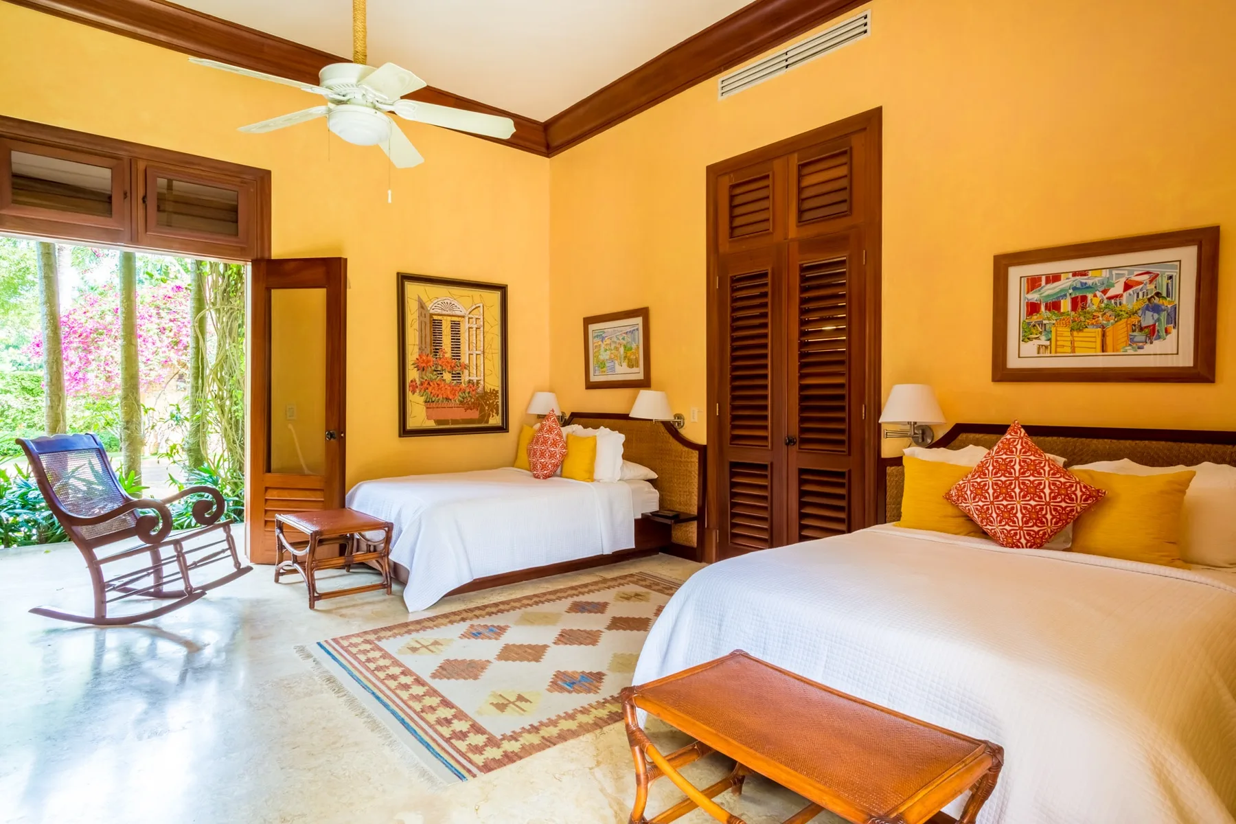 A warmly painted bedroom with two beds, a ceiling fan, and an open doorway to greenery.