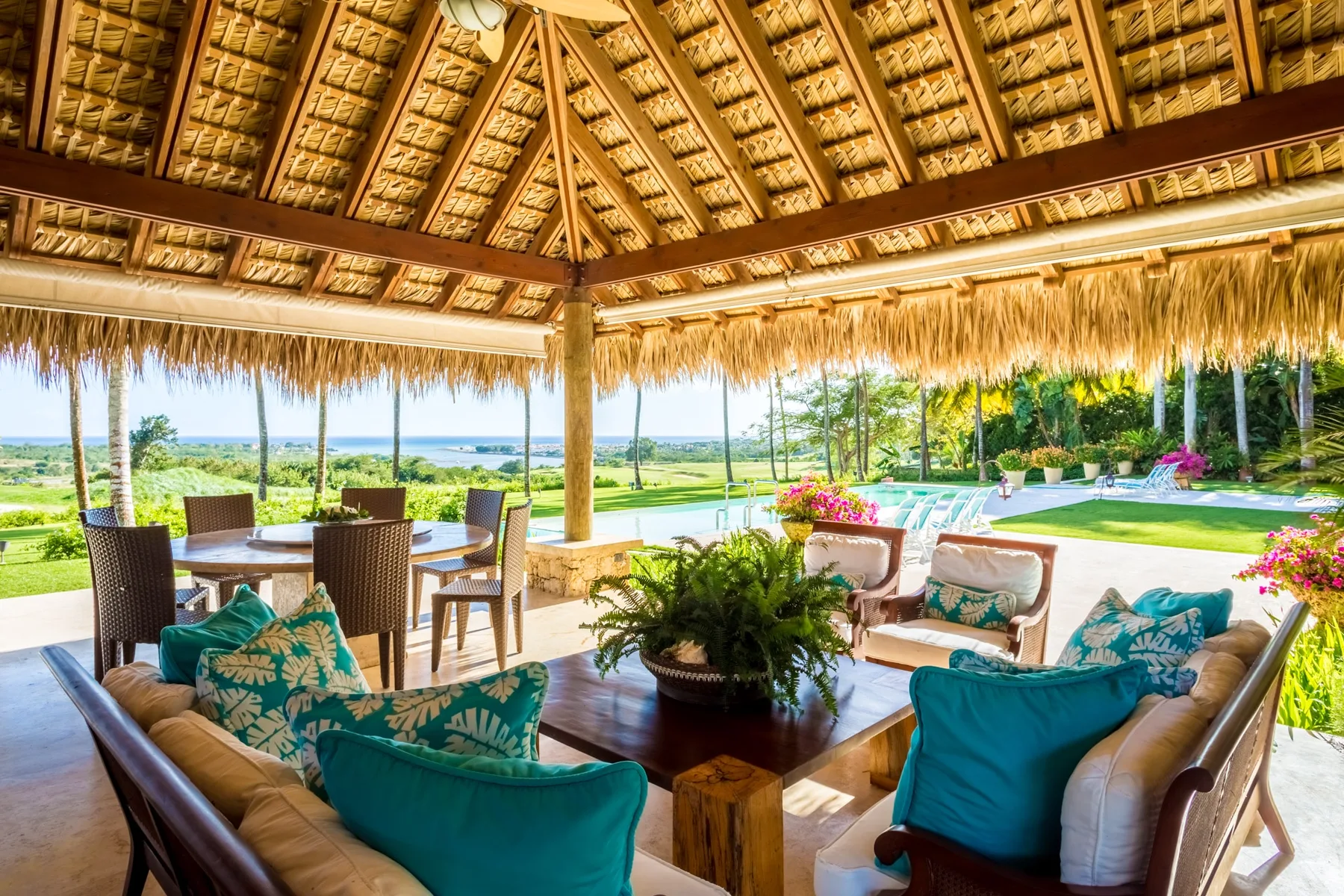 An open-air thatched patio with seating overlooks a garden and swimming pool.