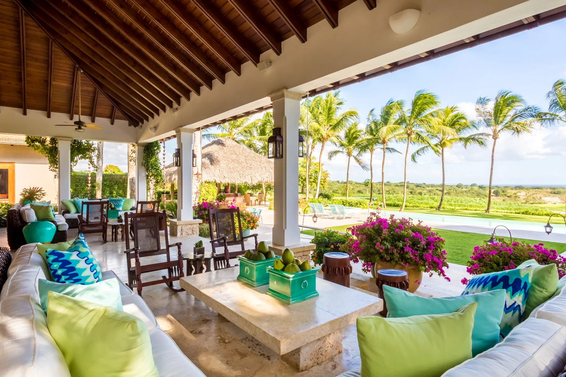 A covered outdoor lounge with cushioned seating overlooks a pool and palm trees.
