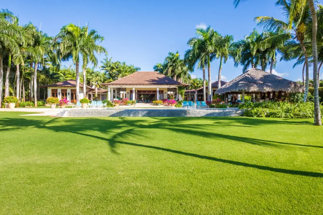 A tropical resort scene with a pool, palm trees, and a broad green lawn.
