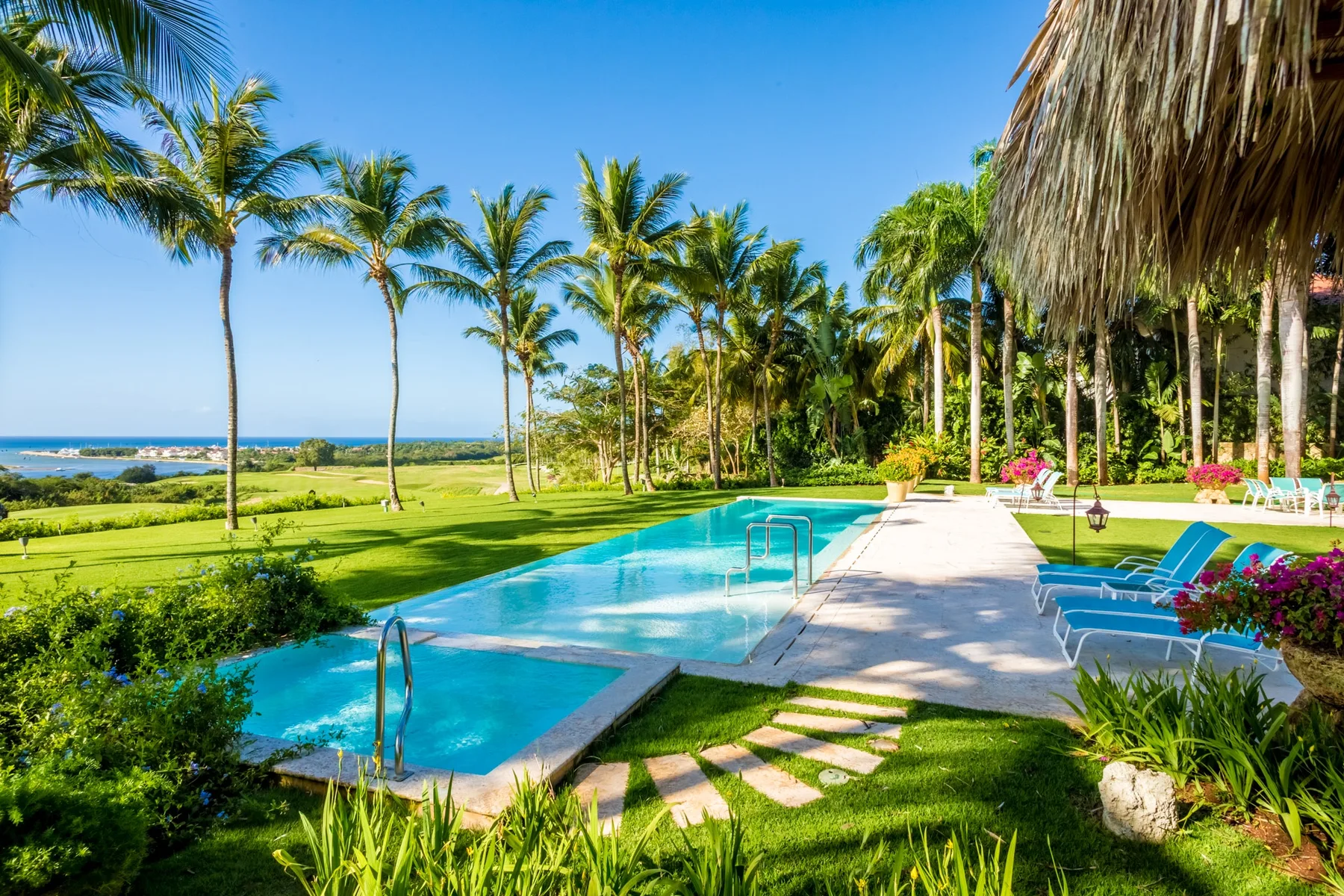 A swimming pool beside a sunlit patio, with palm trees and ocean visible beyond.