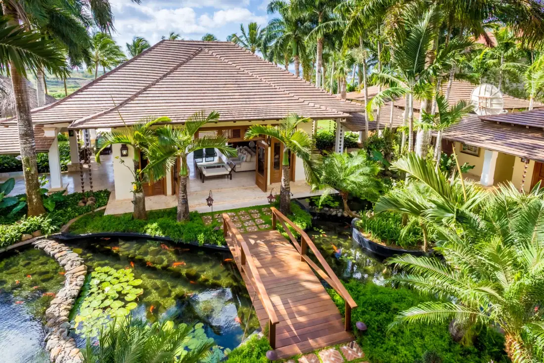 A tropical resort entrance stands beside a garden pond and wooden footbridge.