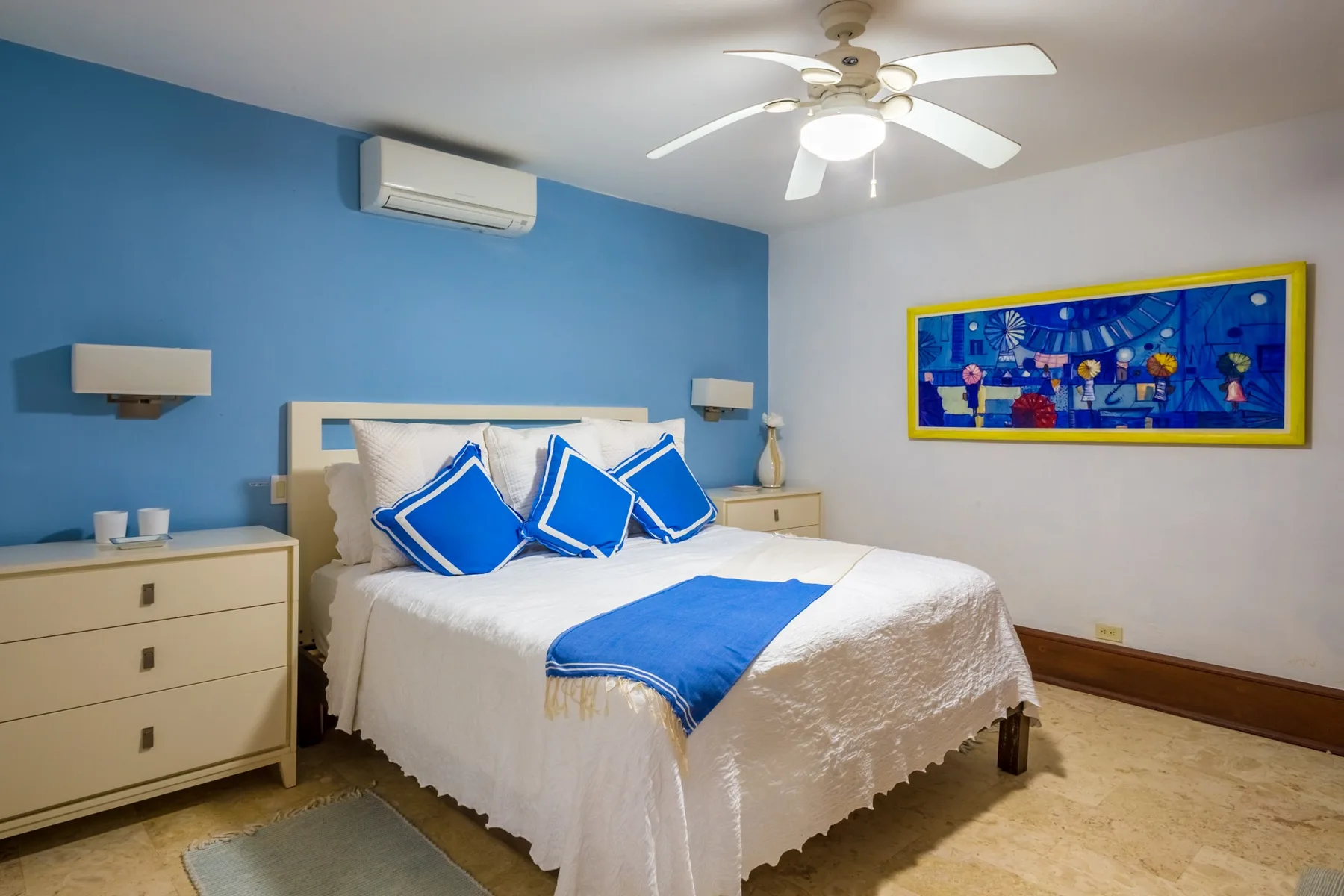 A bedroom with a white bed, blue accent pillows, and colorful wall art.