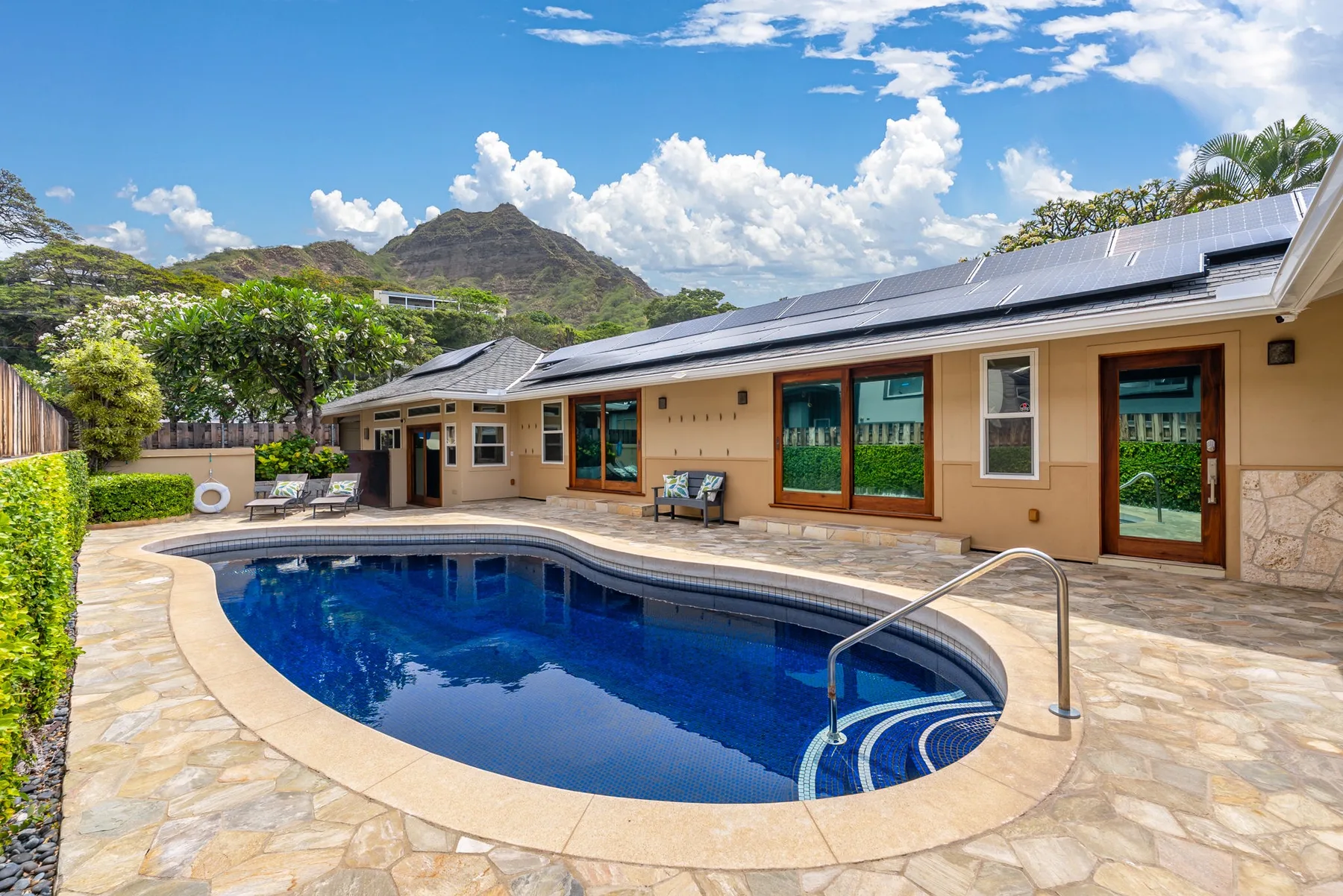 A curved swimming pool sits beside a single-story house with a paved patio.