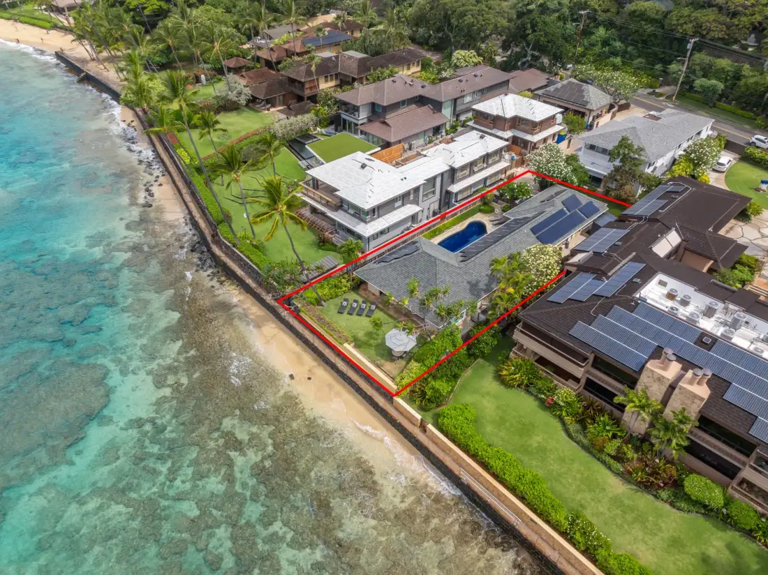 Aerial view of coastal homes, gardens, and a swimming pool beside the shoreline.