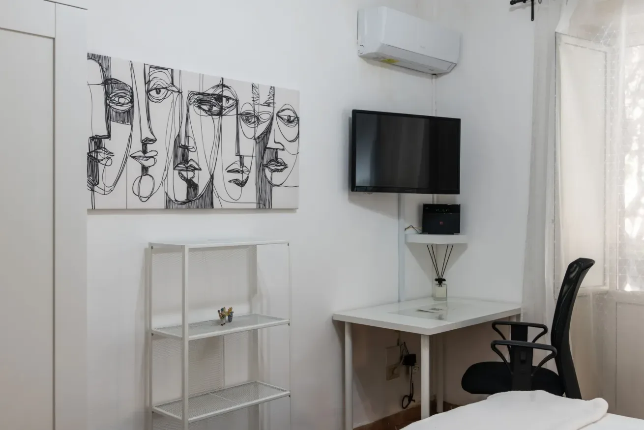 Un espacio de trabajo en un dormitorio con escritorio, televisor en la pared y un cuadro.