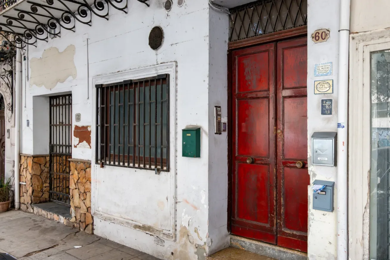 Entrada de un edificio desgastado con una puerta roja y ventanas enrejadas.