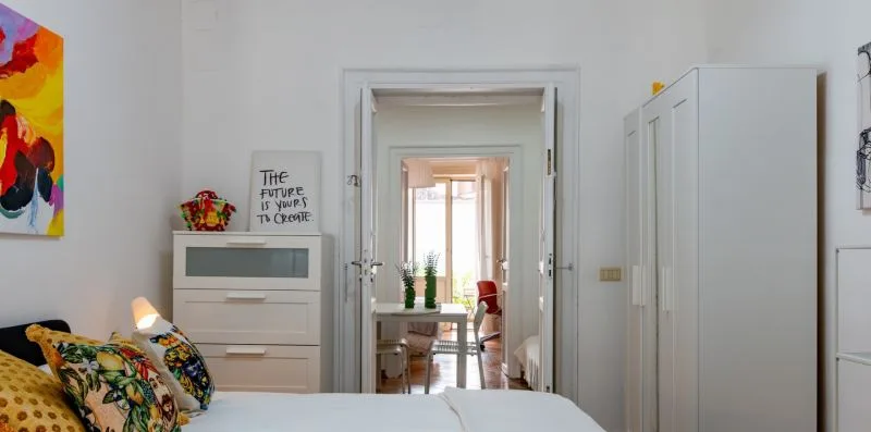 Un dormitorio con una cama, una cómoda y una puerta abierta hacia una habitación contigua luminosa.