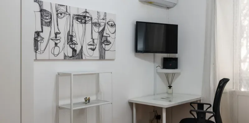 Una habitación luminosa con televisión en la pared, escritorio, silla y arte abstracto de rostros.