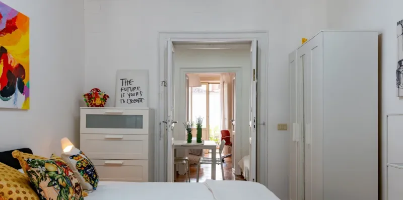 Un dormitorio con una cama, arte colorido en la pared y una puerta hacia una habitación luminosa.