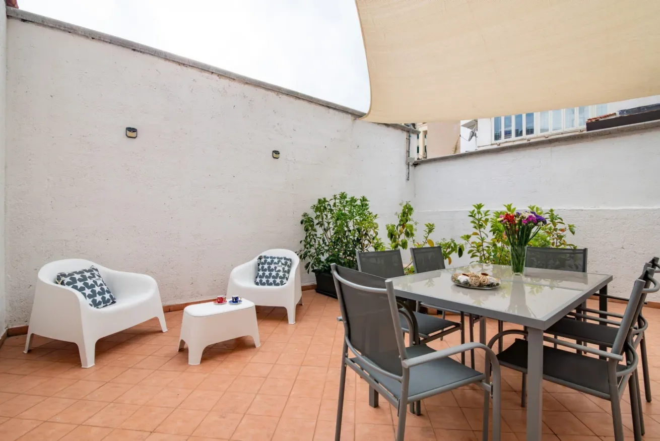 Una terraza exterior con suelo de baldosas, mesa de comedor, asientos y plantas en macetas.
