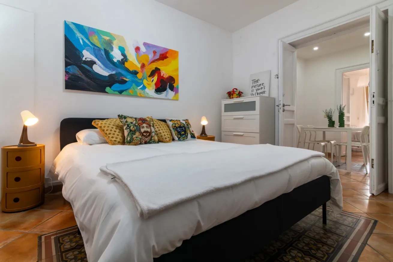 Un dormitorio con una cama bien tendida bajo una colorida pintura abstracta.