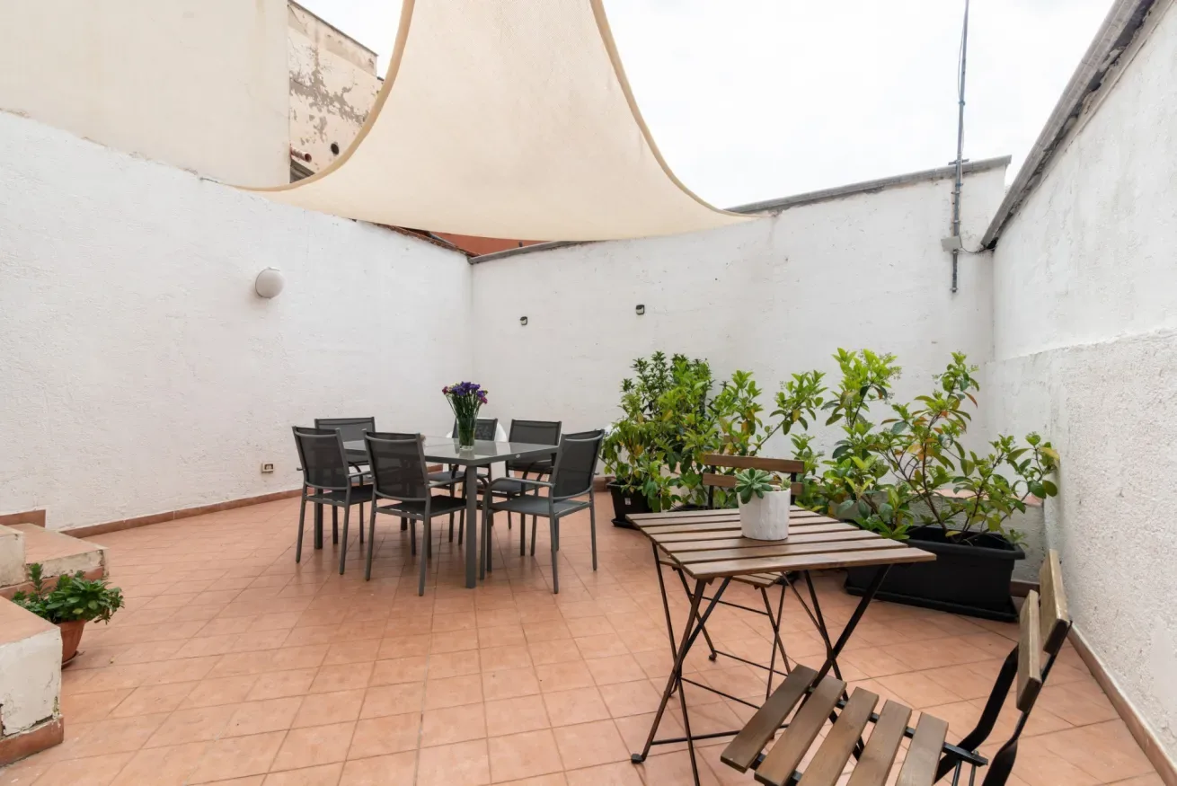 Un patio exterior embaldosado tiene una mesa de comedor, asientos, plantas y un toldo.