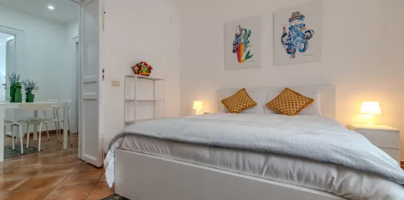 Un dormitorio luminoso con una cama bien tendida y cuadros coloridos encima.