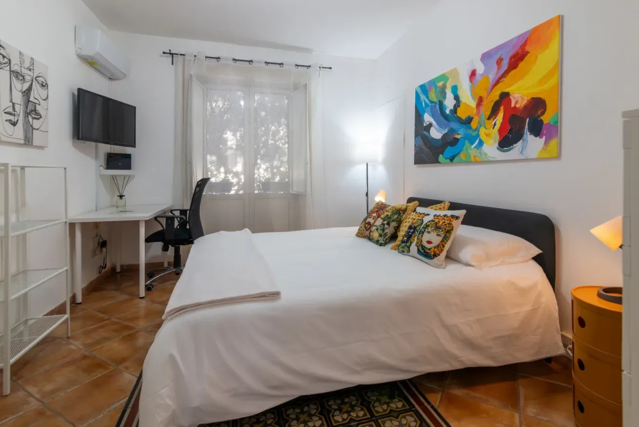 Un dormitorio luminoso con una cama tendida y un escritorio junto a la ventana.