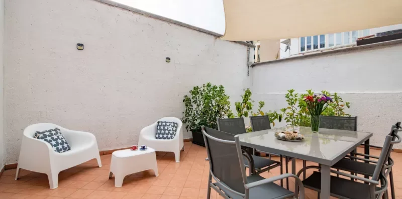 Una zona de comedor al aire libre tiene una mesa de cristal, asientos y plantas en macetas.