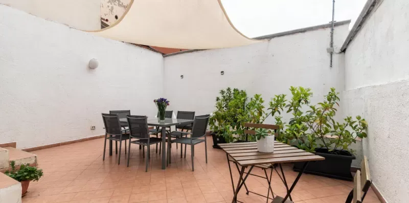 Un patio con suelo de baldosas tiene una mesa, asientos y plantas en macetas.