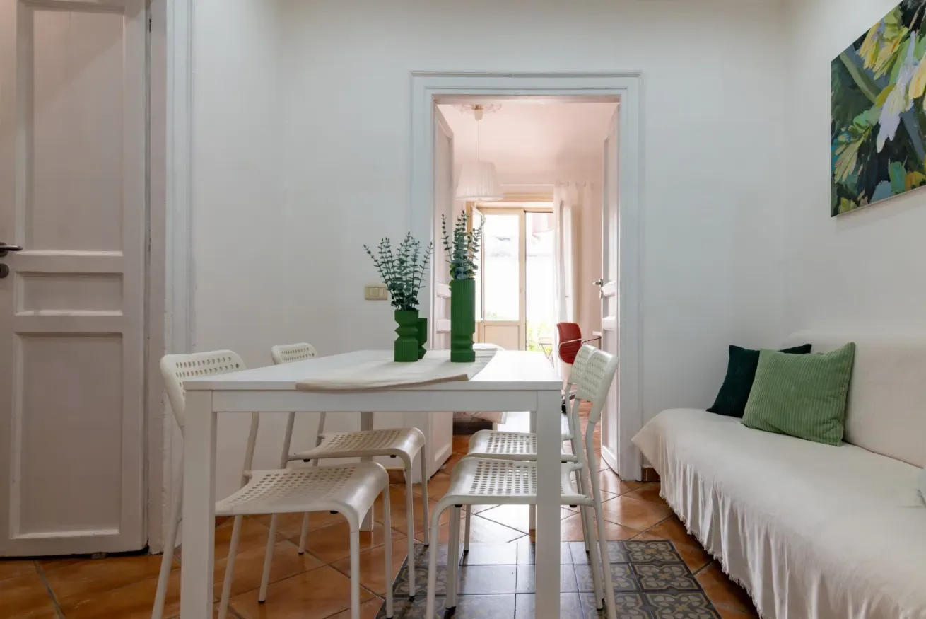 Una mesa de comedor con sillas está junto a un sofá en una sala luminosa.