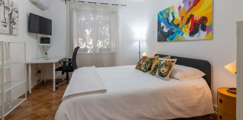 Un dormitorio con una cama tendida, arte colorido en la pared y un escritorio junto a la ventana.
