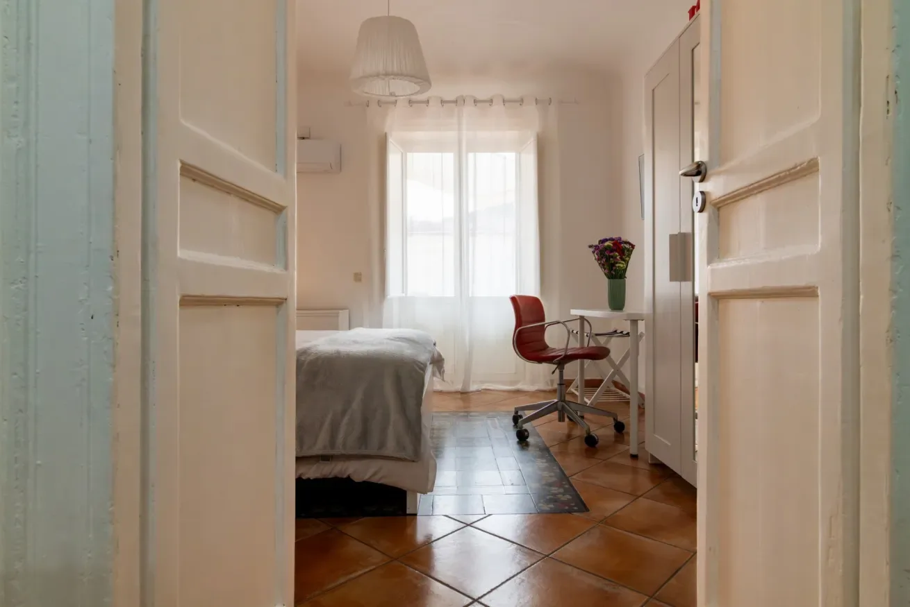 Un dormitorio luminoso con una cama, un escritorio y una silla roja junto a la ventana.