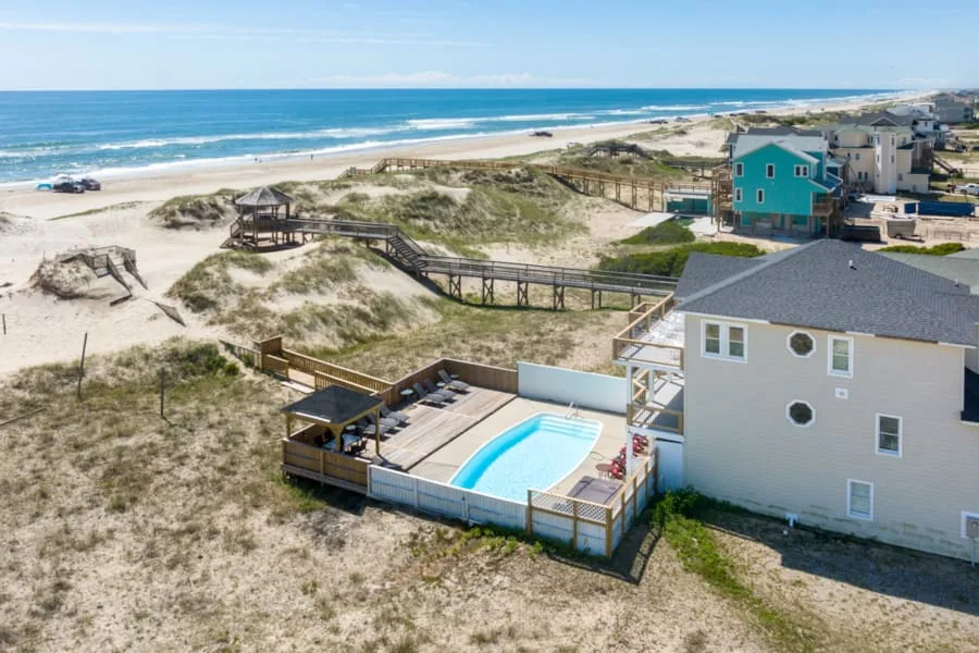 A coastal house with a pool deck sits beside sandy dunes and the ocean.