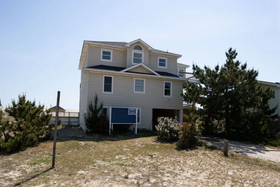 A large light-colored house stands among shrubs beside a sandy yard.