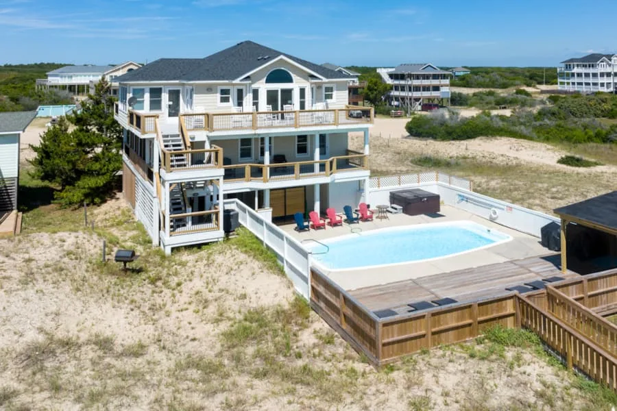 A large coastal house overlooks an outdoor swimming pool and surrounding sandy terrain.