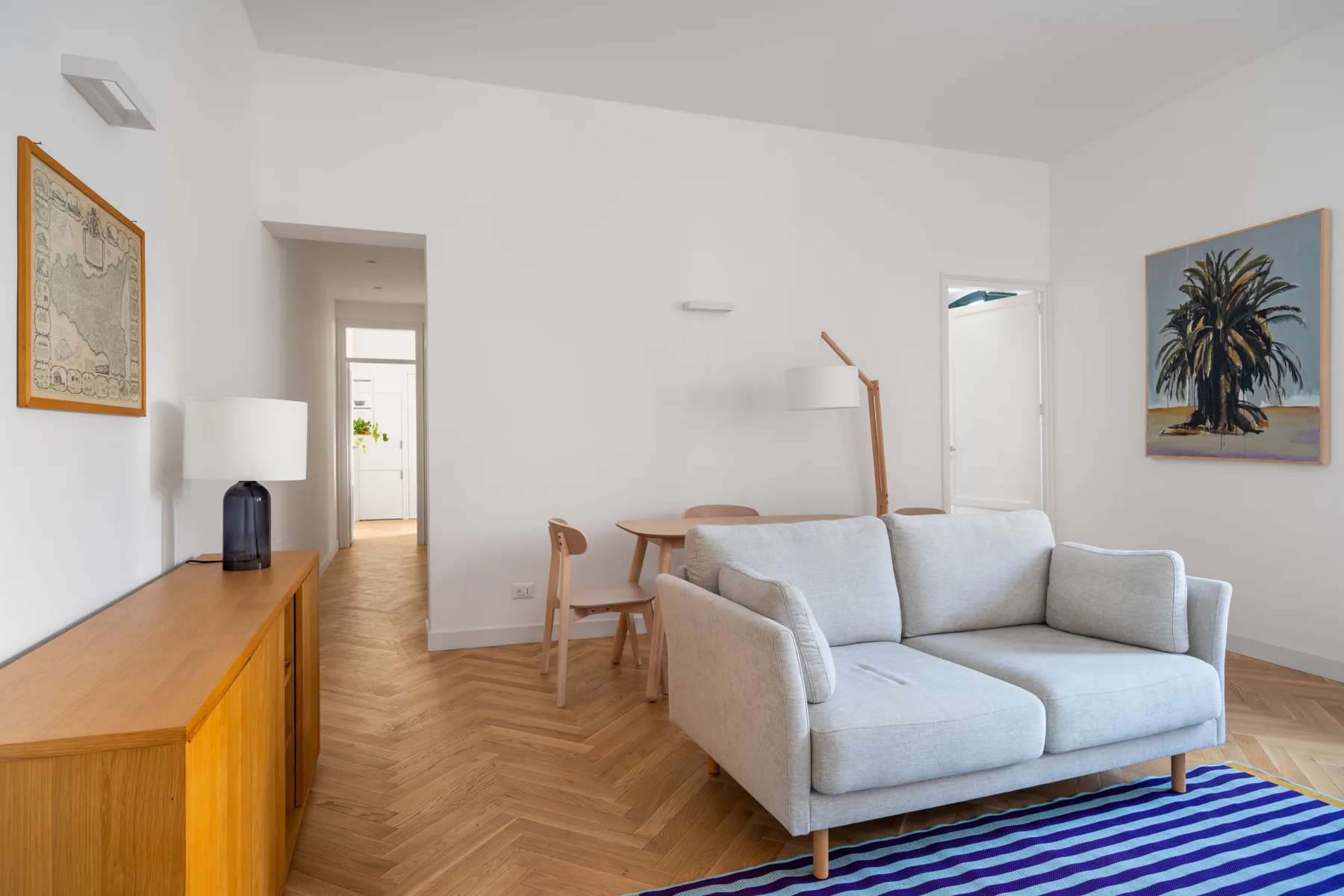 A bright living room with light seating, a wooden sideboard, and framed artwork.