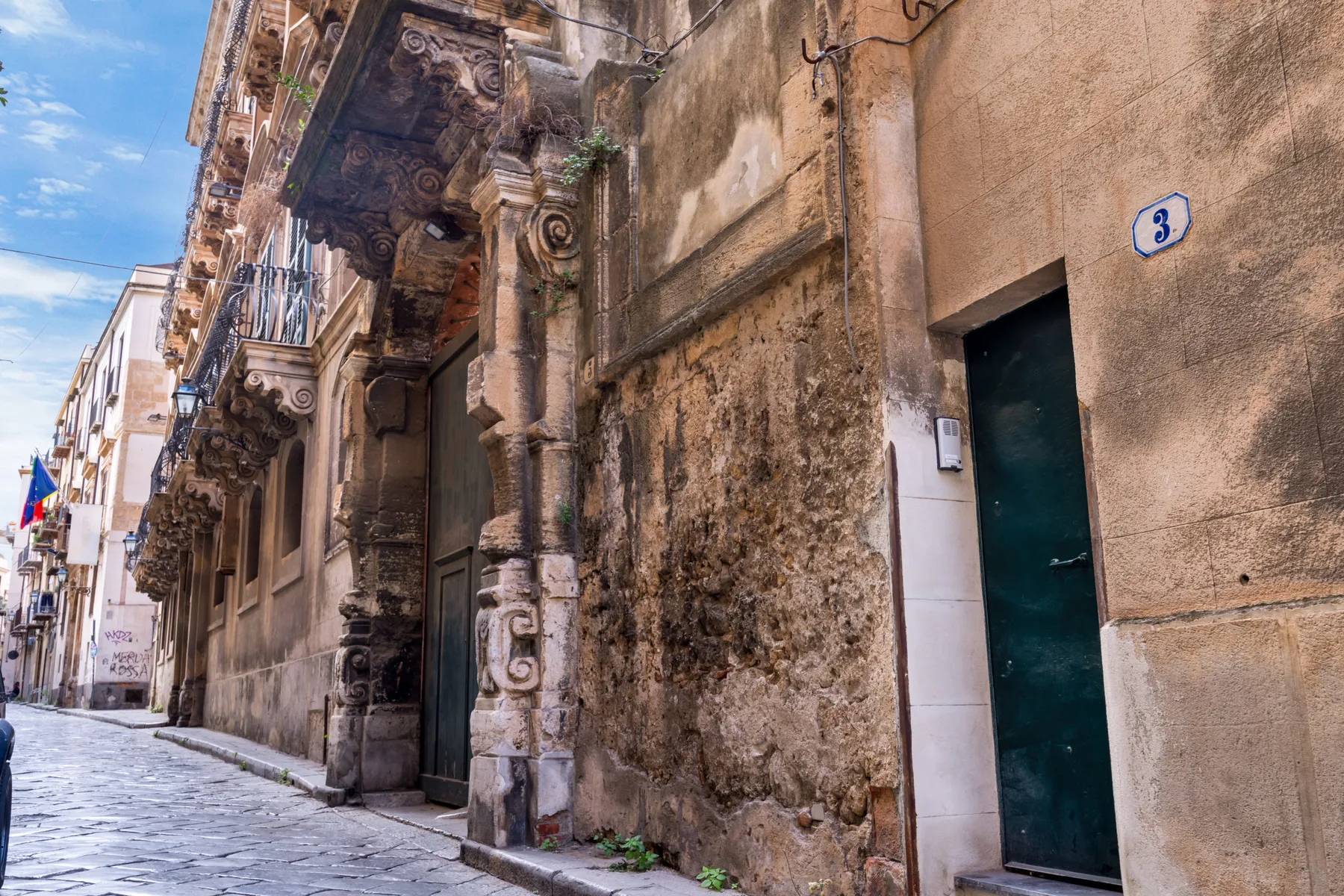 A narrow cobblestone street lined with weathered stone buildings and ornate balconies.