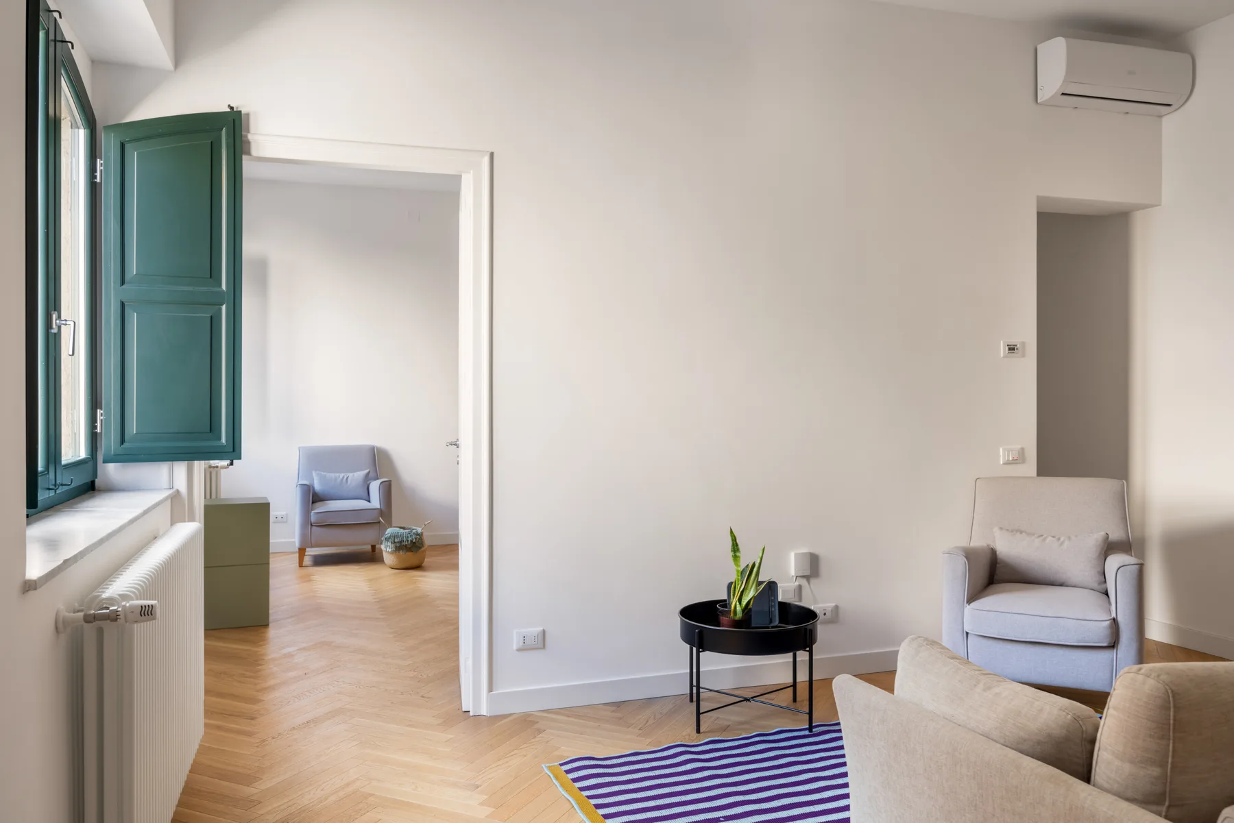 A bright living room with pale seating, a small table, and a green-shuttered window.