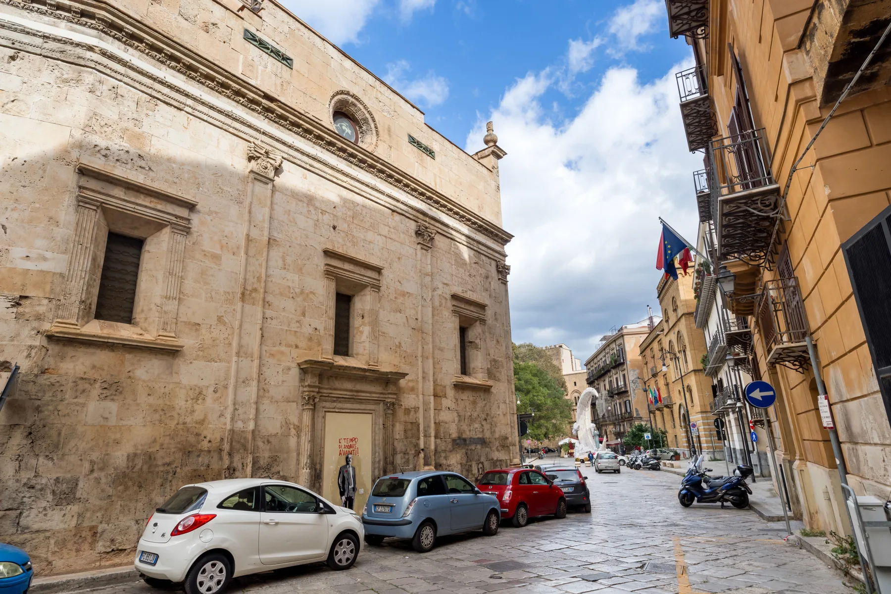 A historic stone building faces a narrow city street lined with parked cars.