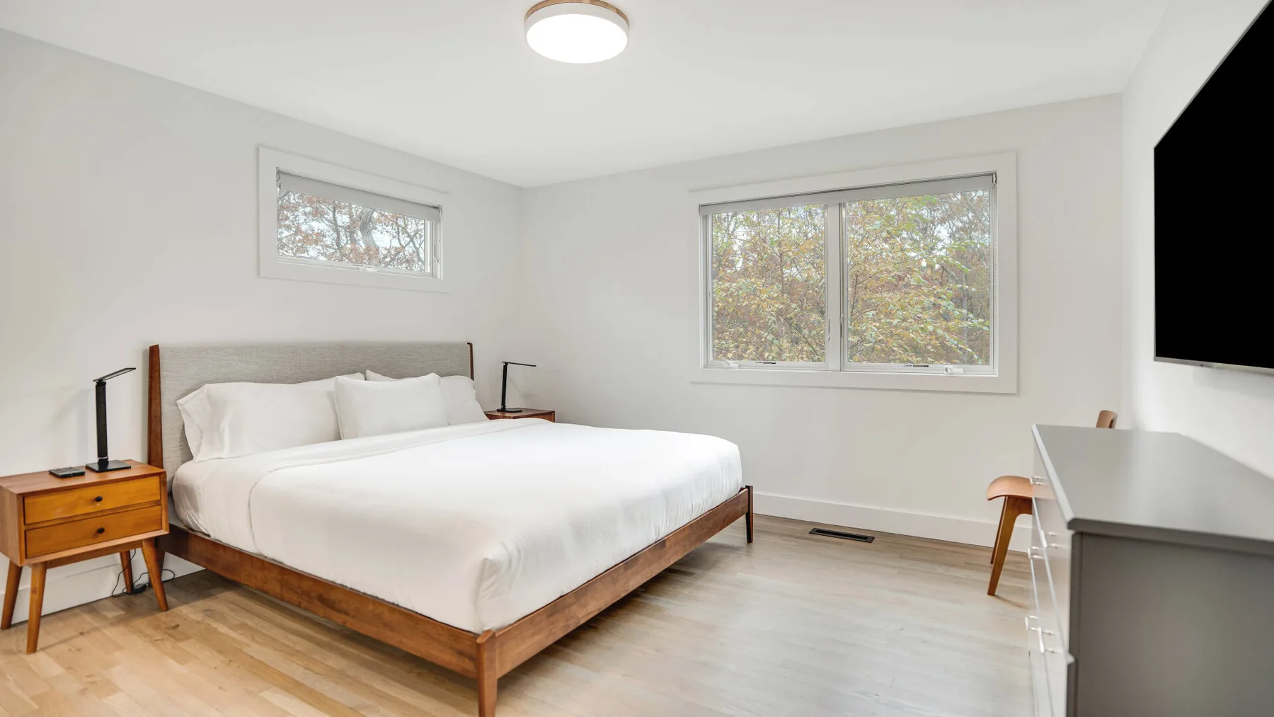 Un dormitorio luminoso con una cama tendida y ventanas con vistas a los árboles.