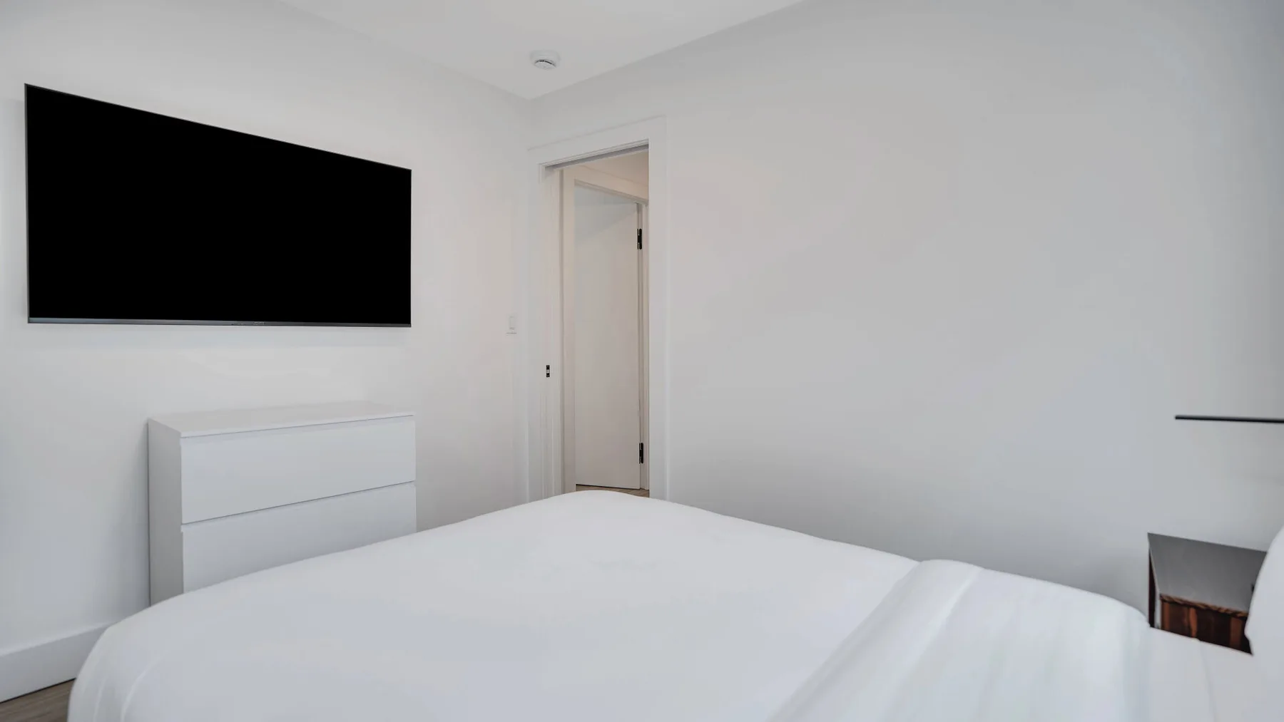 Un dormitorio luminoso con una cama tendida y un televisor montado en la pared.