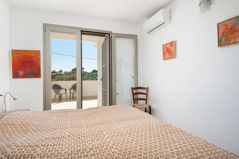 Un dormitorio con una colcha estampada, puertas abiertas al balcón y una silla.