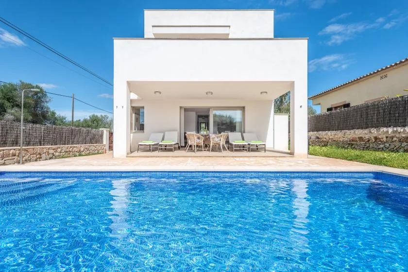 Una casa blanca moderna da a una piscina azul, con asientos al aire libre bajo un patio cubierto.