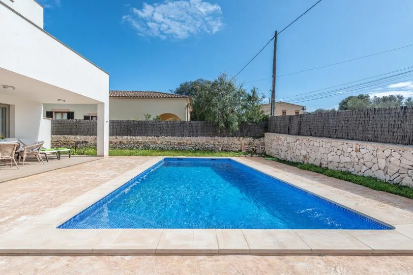 Una piscina rectangular al aire libre junto a un edificio blanco y un patio pavimentado.