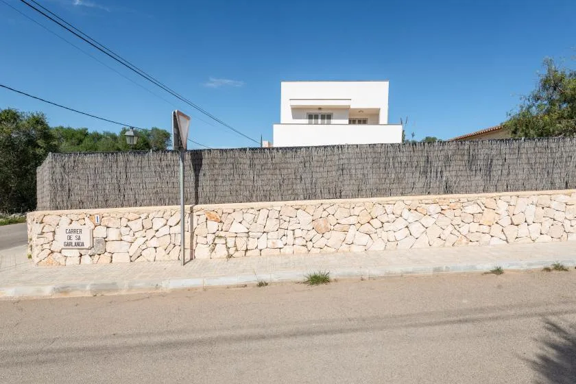 Un edificio blanco se alza detrás de un muro de piedra y una cerca alta junto a la calle.