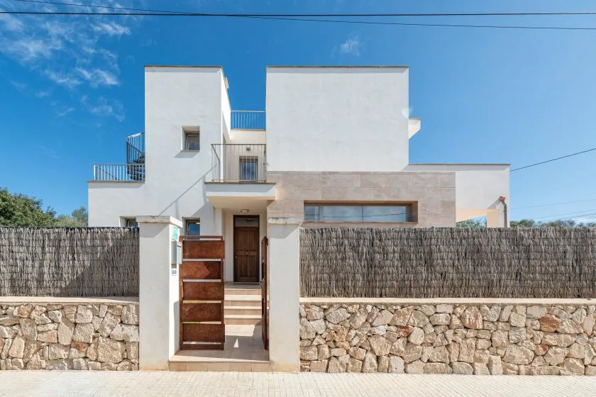 Una casa blanca moderna se alza detrás de un muro de piedra y una puerta de madera.