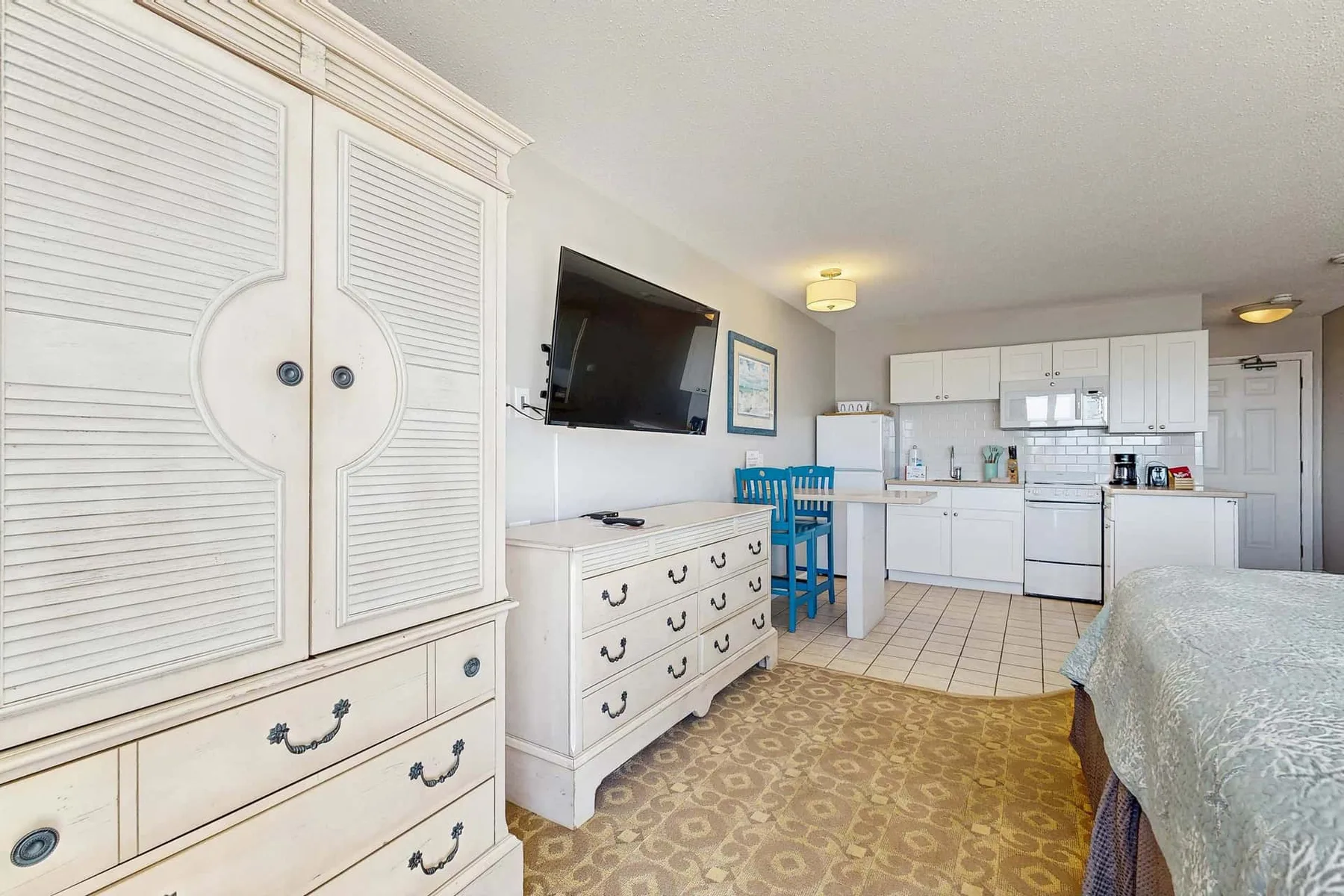 A bedroom with a kitchenette, featuring a bed, white cabinets, and a small dining table.