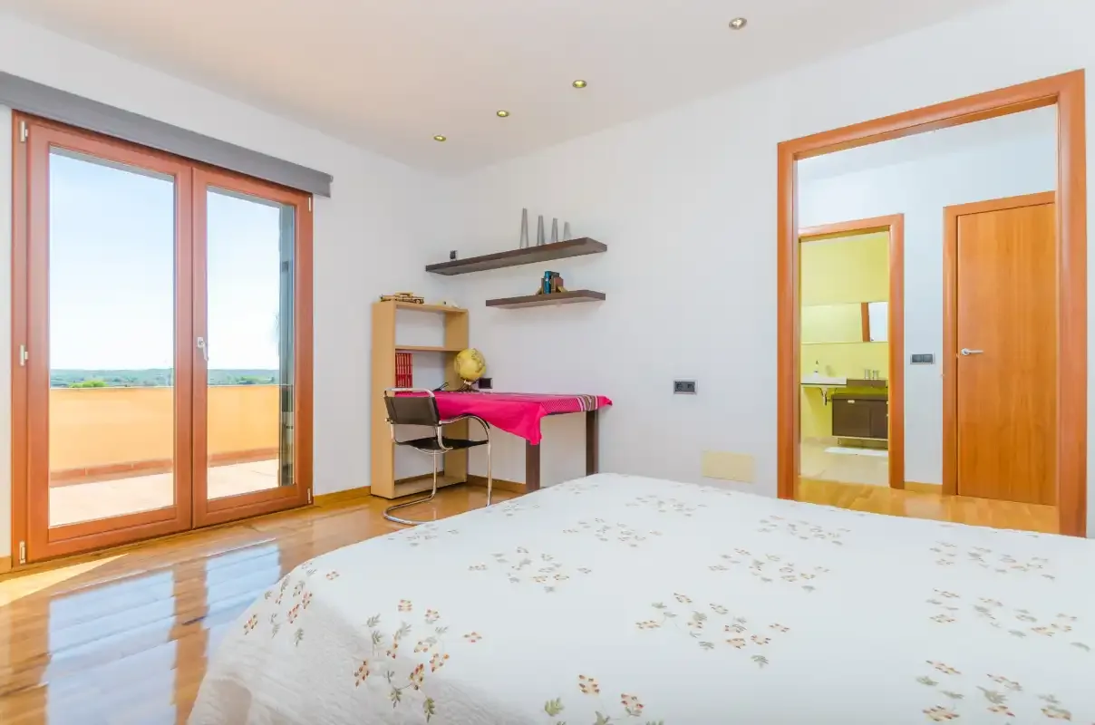 Un dormitorio con una cama, un pequeño escritorio y puertas de vidrio que dan a un balcón.