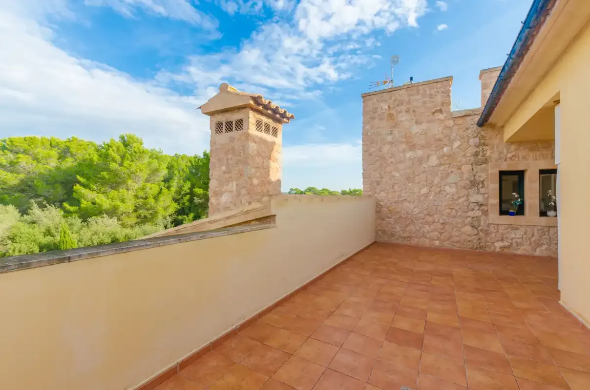 Una terraza exterior con suelo de baldosas da a árboles verdes junto a un edificio de piedra.